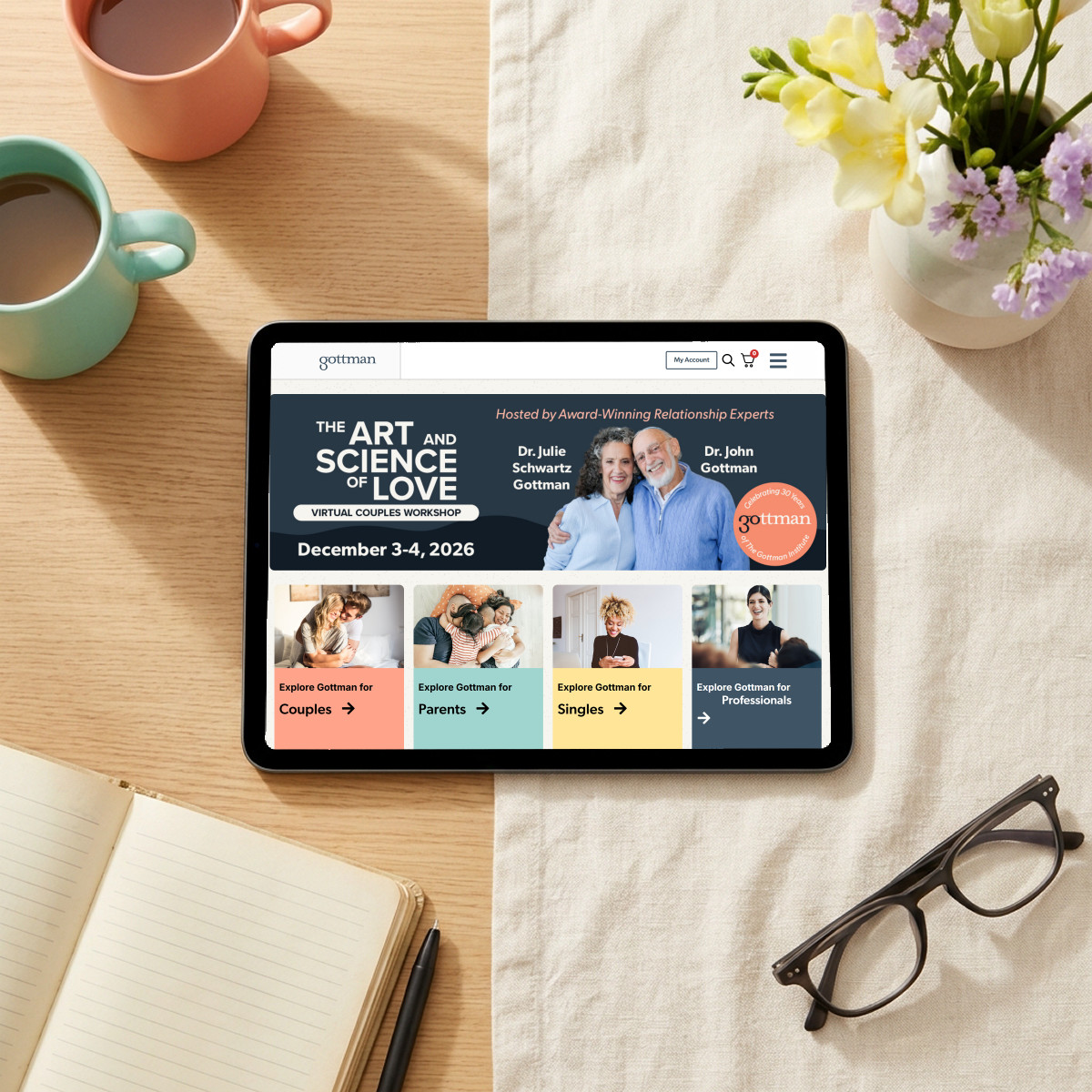 Gottman Institute website on a tablet, beside two coffee mugs, flowers and an open journal